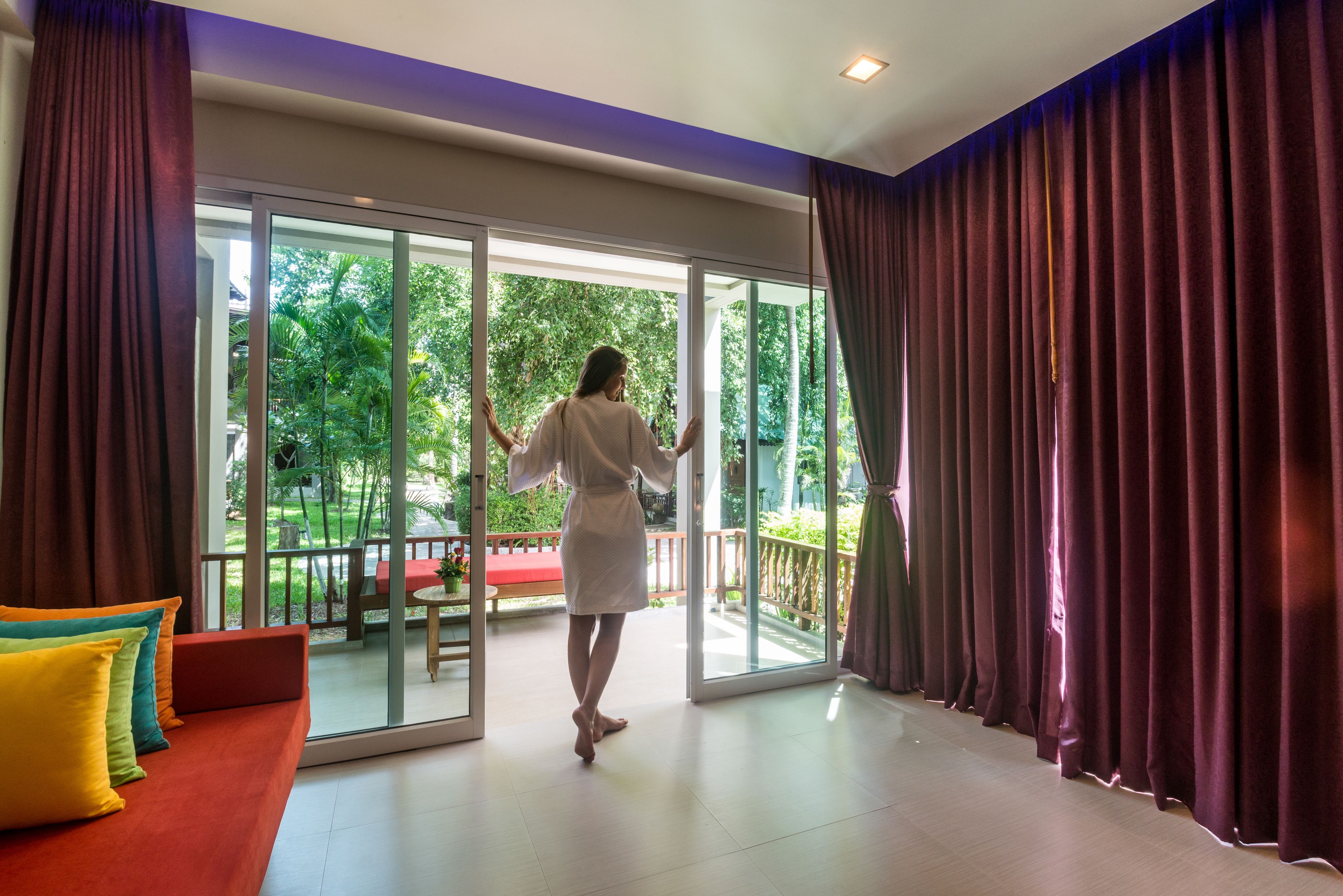 A room with a sliding glass door leading to a balcony with a red bench and greenery.