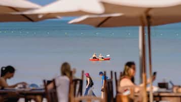 Een strandscène met een rode kajak in het water, mensen die langs het strand lopen, en parasols die schaduw bieden.