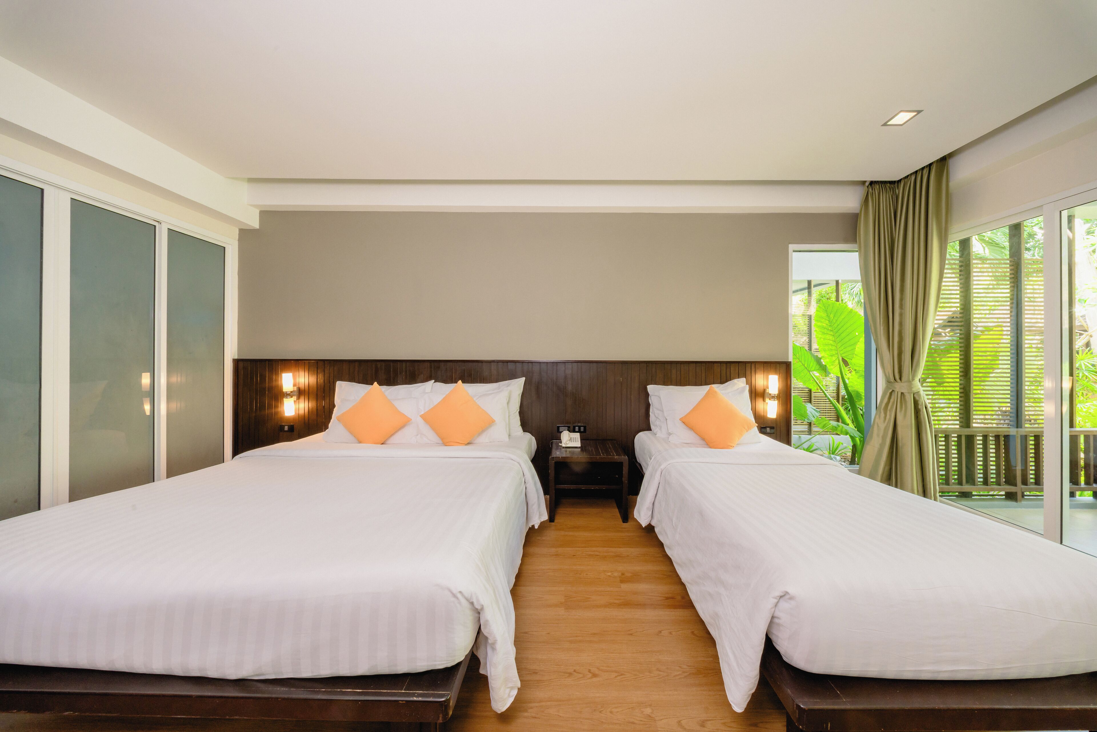 A hotel room with two beds, wooden flooring, a nightstand, and a view of greenery through large windows.