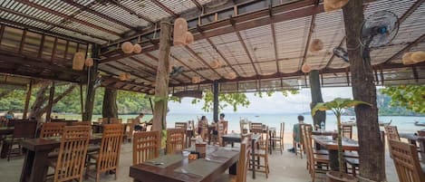 Un restaurante al aire libre con mesas y sillas de madera, techo metálico y vista al mar.