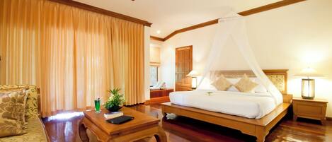 Una habitación de hotel con una cama de madera, una mesita de noche de madera, una mesa de centro de madera, un sofá y un ventanal grande con cortinas.