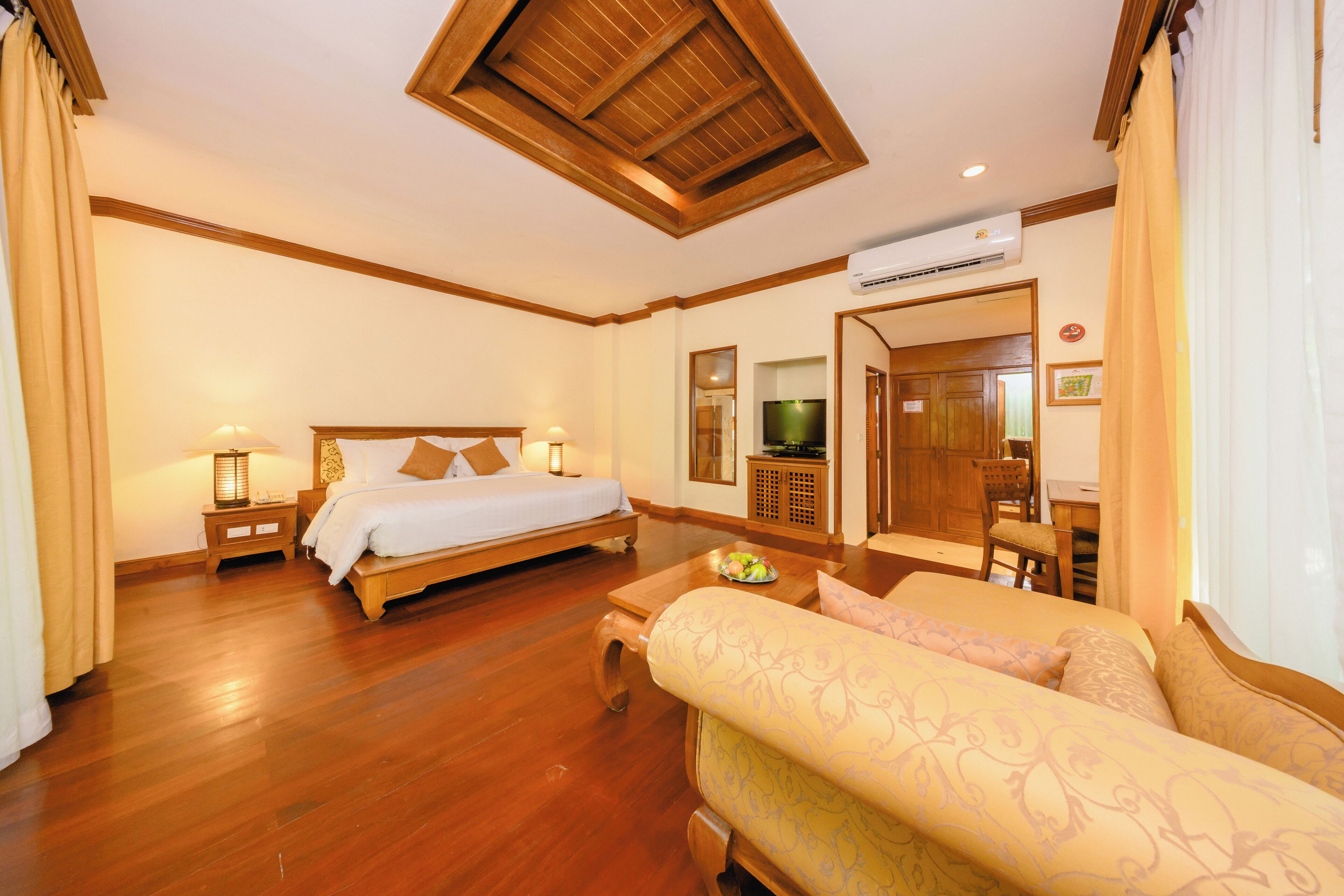 A hotel room with a wooden floor, a bed with a white comforter, a sofa, a small table with a fruit bowl, a television, and a wooden cabinet.