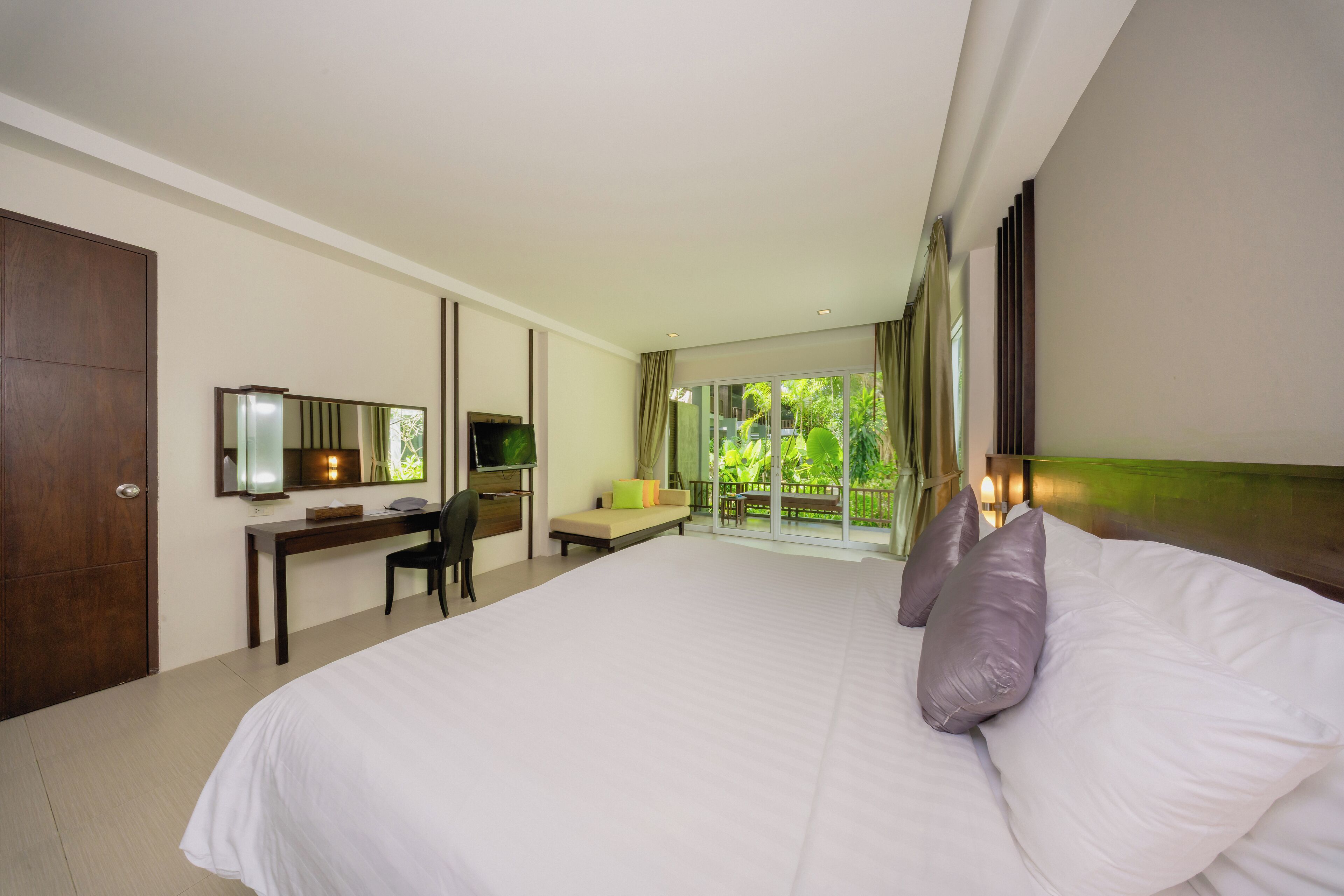 A hotel room with a large bed, a desk with a chair, a television, and a balcony with a view of greenery.