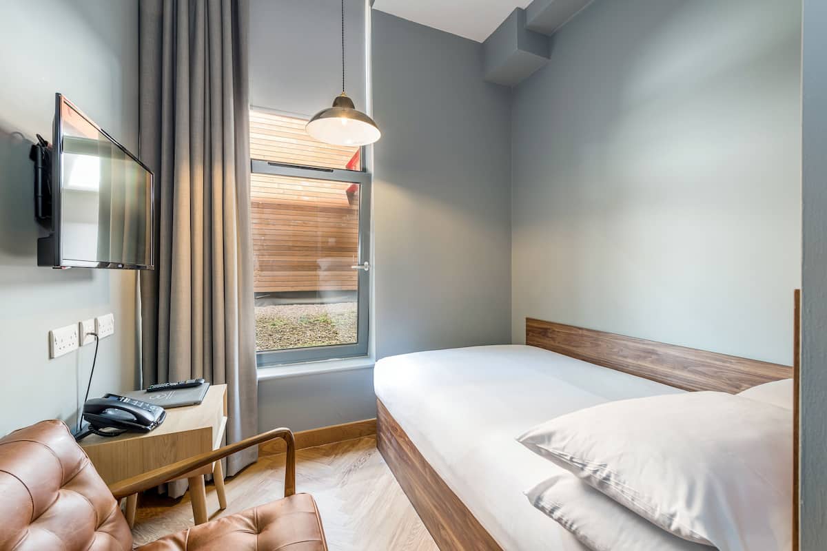 Une chambre d’hôtel moderne équipée d’un lit, d’un bureau avec un ordinateur portable, d’un téléphone et d’un téléviseur fixé au mur.