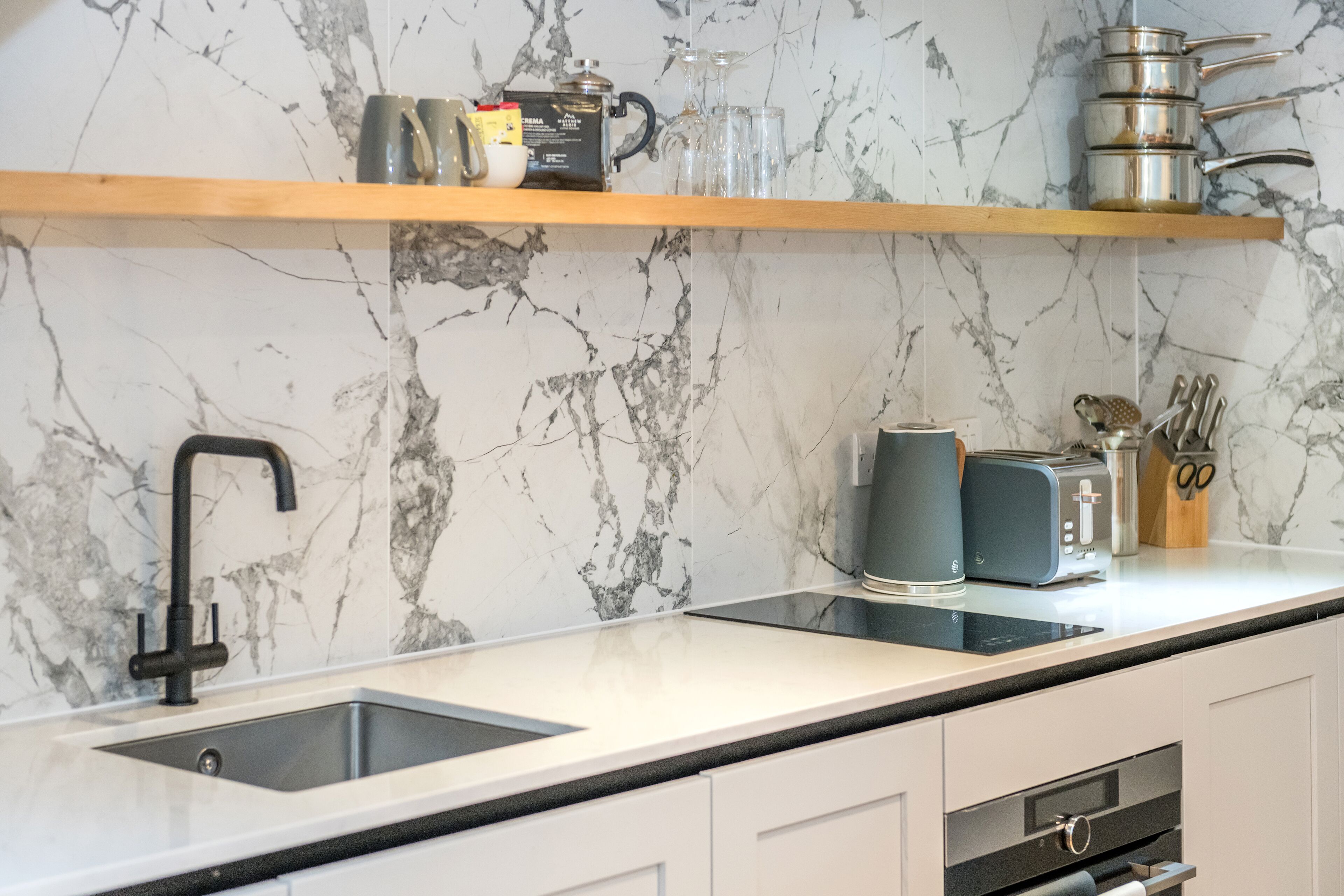 A modern kitchen with a marble backsplash, a black faucet, a built-in oven, and a wooden shelf above the sink.
