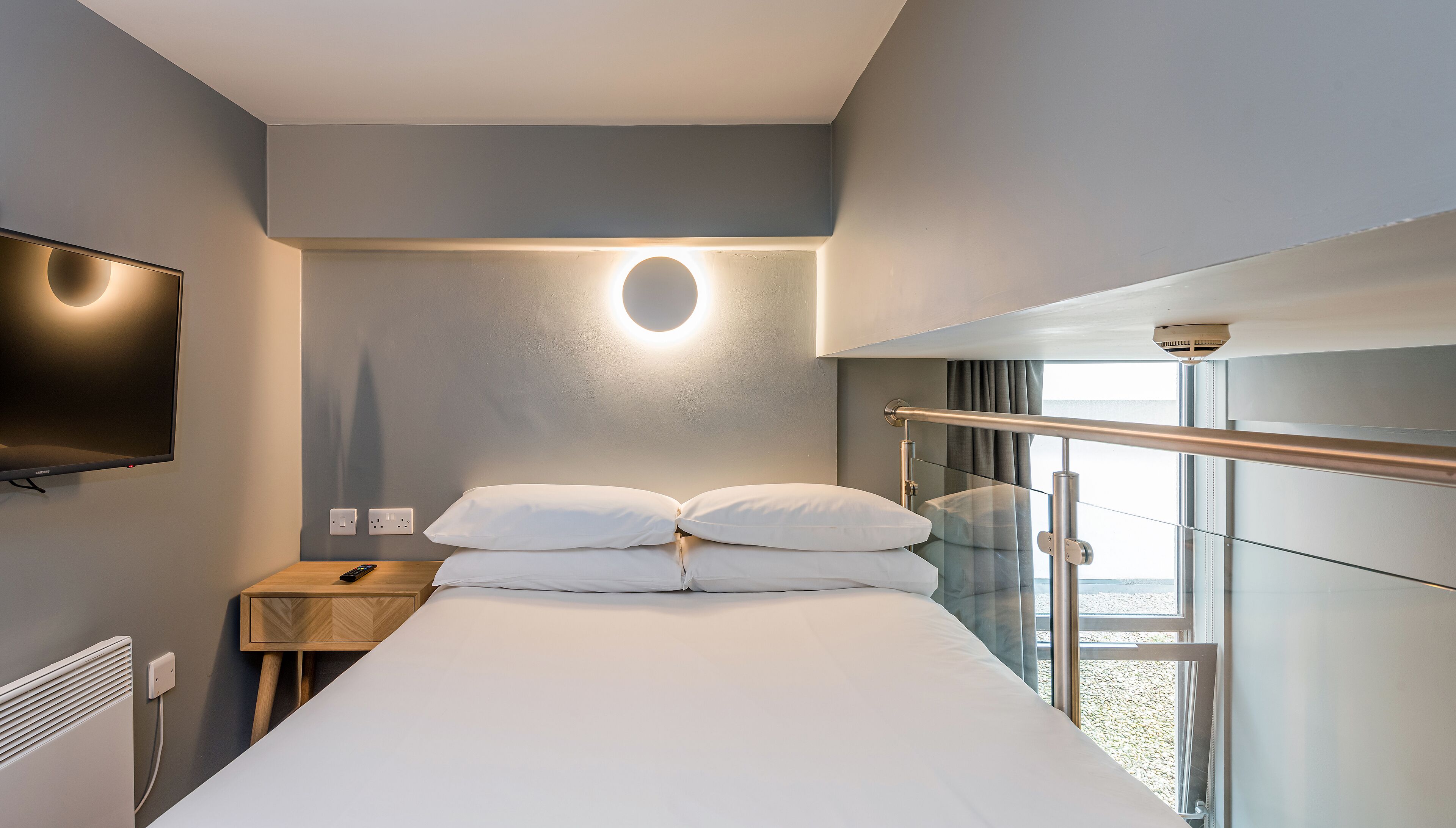 Una habitación de hotel moderna con cama, televisor de pantalla plana, mesita de noche, ventana con vistas y separador de vidrio.
