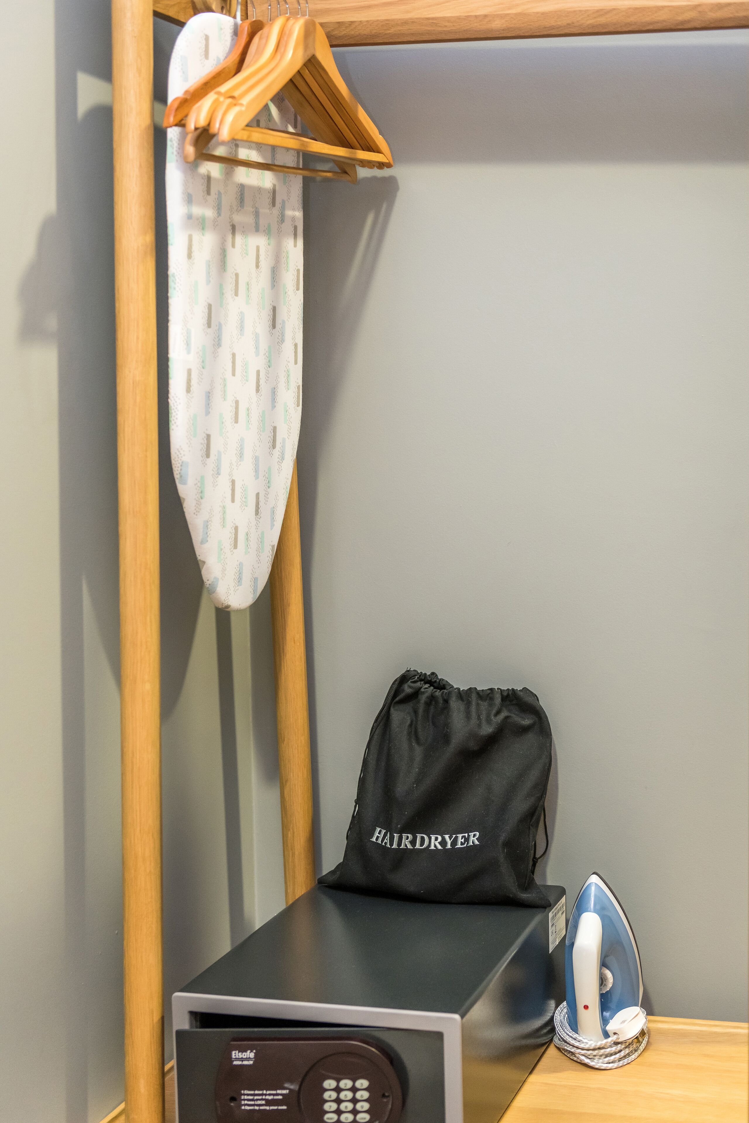 Habitación de hotel con secador de pelo, plancha y una bolsa negra con la etiqueta 'HAIRDRYER' sobre una mesa de madera.
