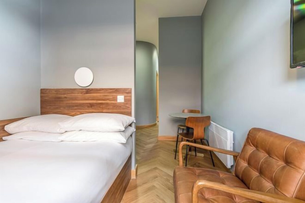 A modern hotel room with a wooden headboard bed, a round mirror, a brown leather sofa, and a small table with chairs.