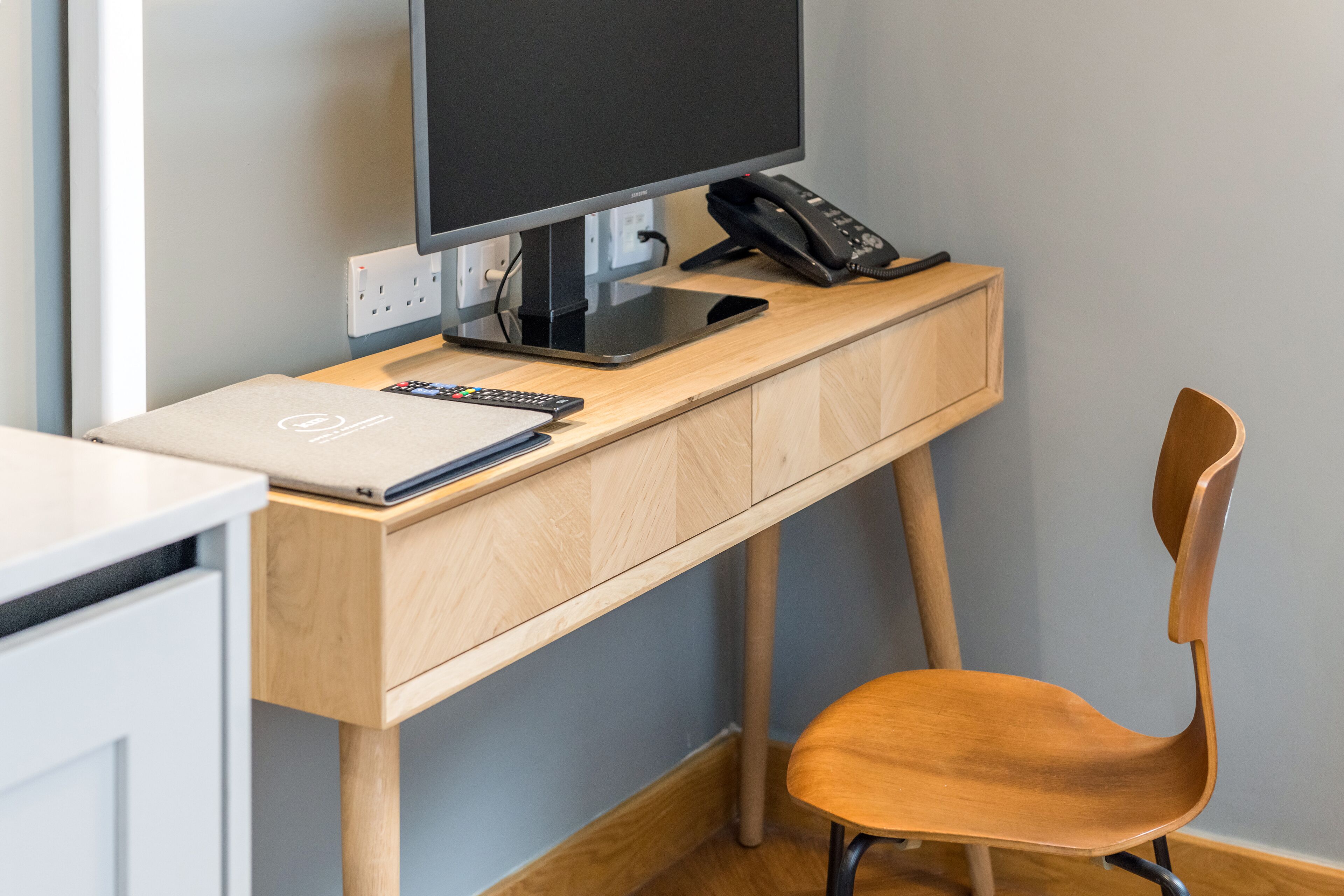A wooden desk with a computer monitor, phone, and remote control. A chair is positioned in front of the desk.