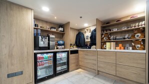Una kitchenette moderna con cafetera, dos mini refrigeradores llenos de bebidas, y estantes con aperitivos y artículos de tocador.