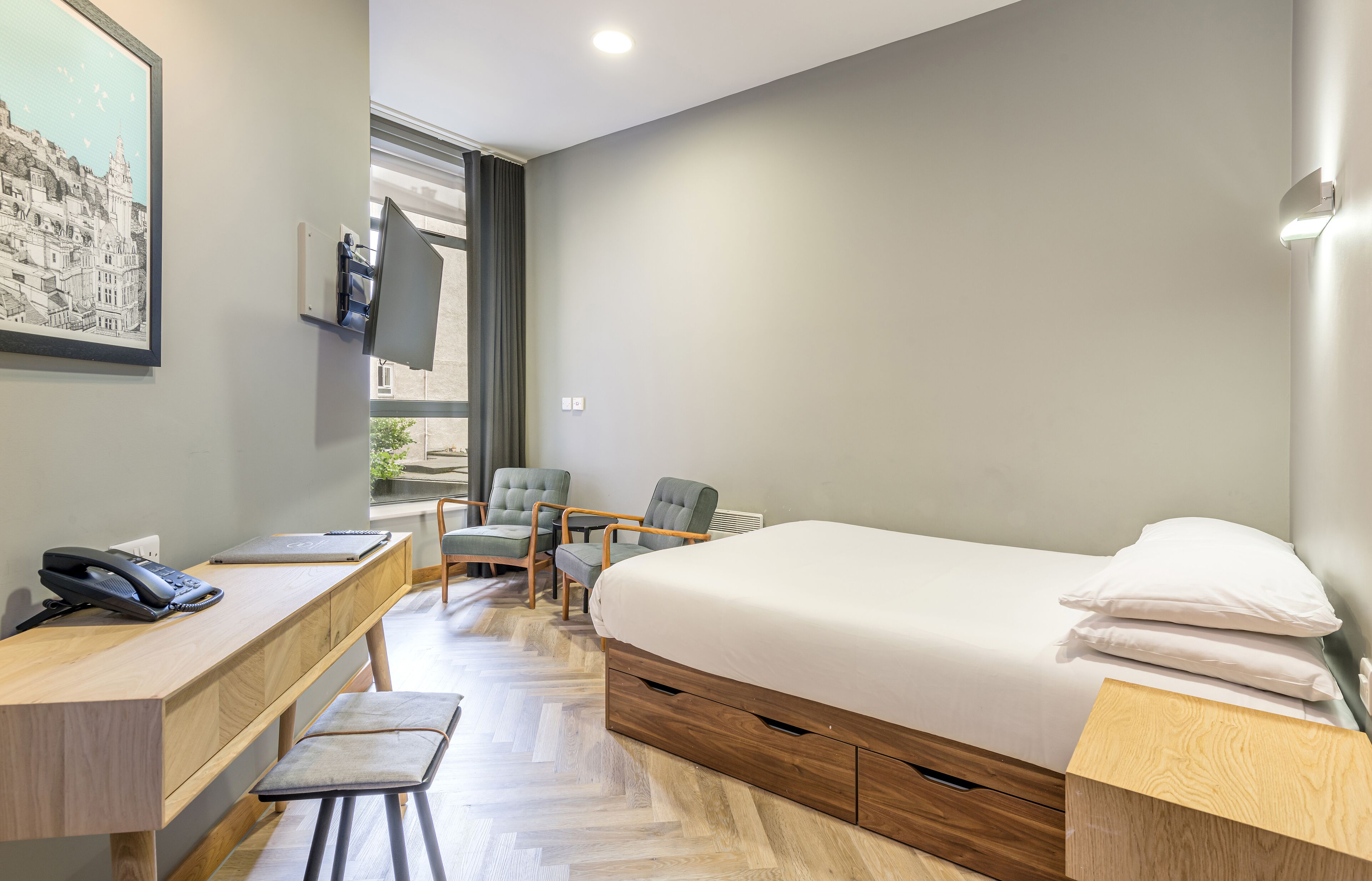 A hotel room with a bed, desk, chairs, a phone, and a window.