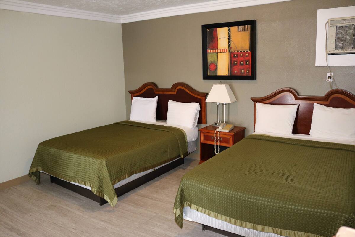 Ein Hotelzimmer mit zwei Betten, jedes mit einem hölzernen Kopfteil, einer grünen Bettdecke und weißen Kissen. Zwischen den Betten befindet sich ein Nachttisch mit einer Lampe und einem Telefon.