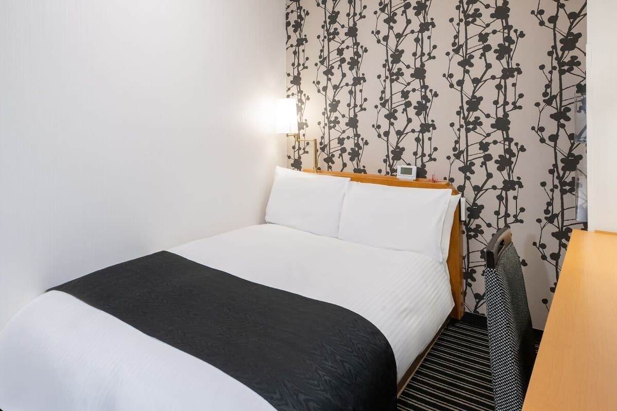 Una cama bien tendida con una colcha blanco y negro, cabecera de madera y pared con estampado floral negro.