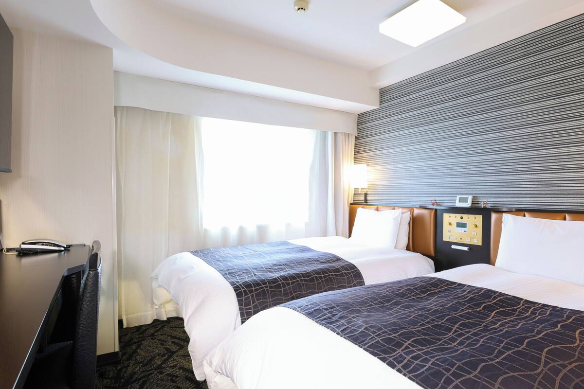A hotel room with two beds, a desk, a telephone, and a window with curtains.