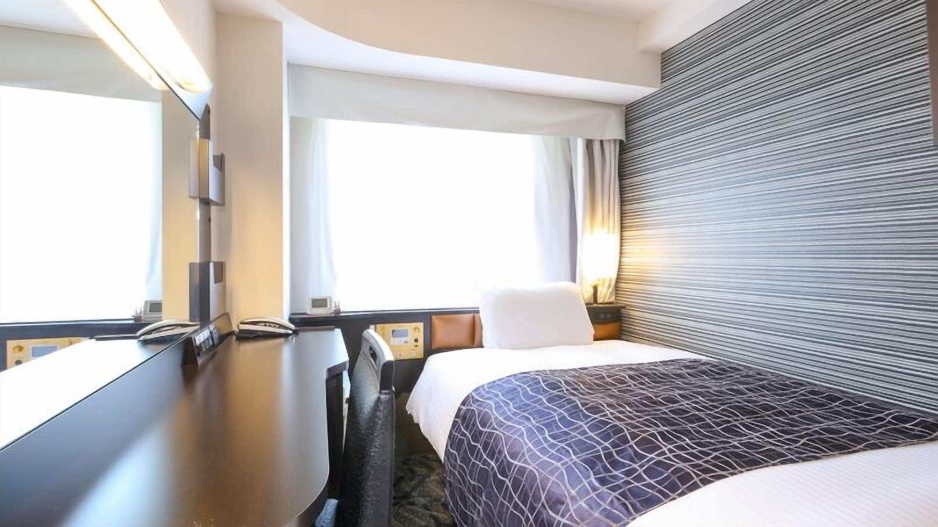 Ein Hotelzimmer mit Bett, Schreibtisch, Stuhl, Nachttisch und einem Fenster mit Vorhängen.