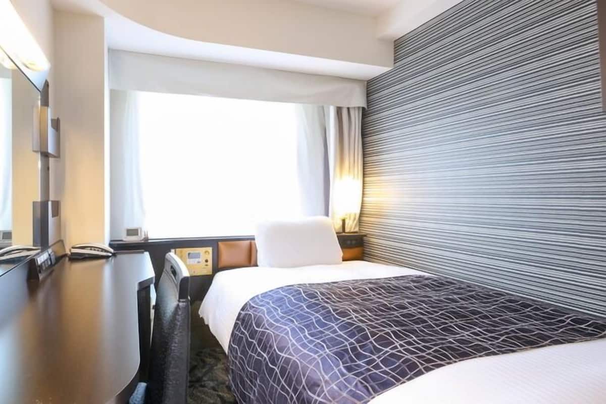 A hotel room with a bed, a desk, a chair, a bedside table, and a window with curtains.