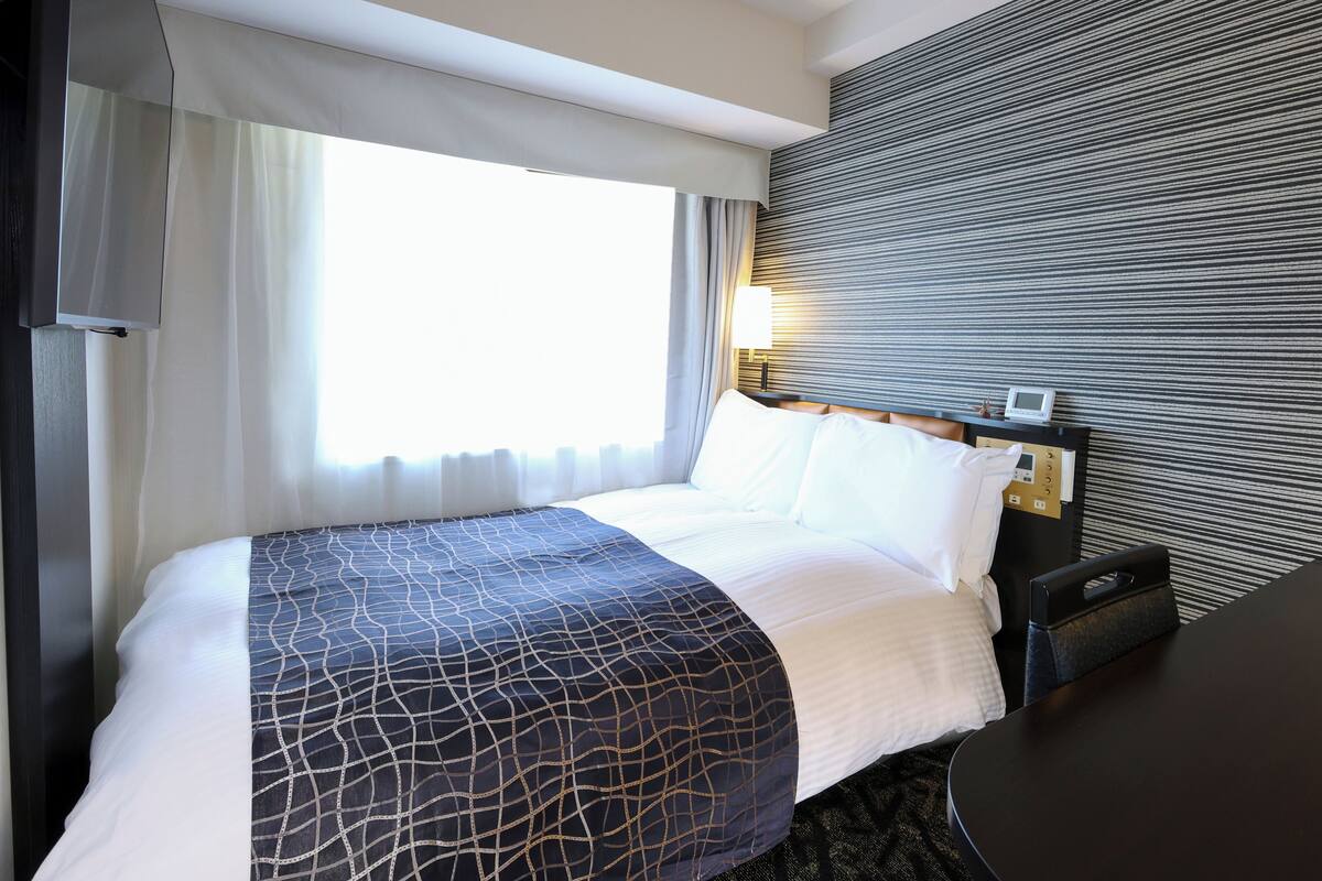 A hotel room with a bed, a bedside table with a lamp, and a view of the outdoors through a window with sheer curtains.