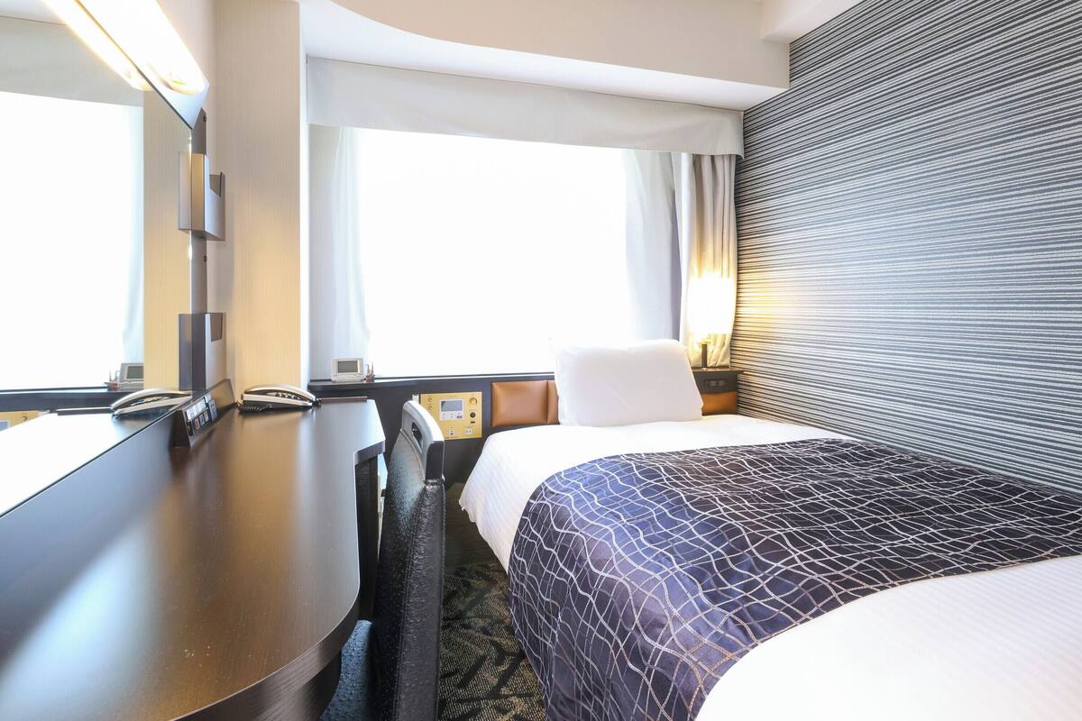 A hotel room with a bed, a desk, a chair, a bedside table, a lamp, and a window with curtains.