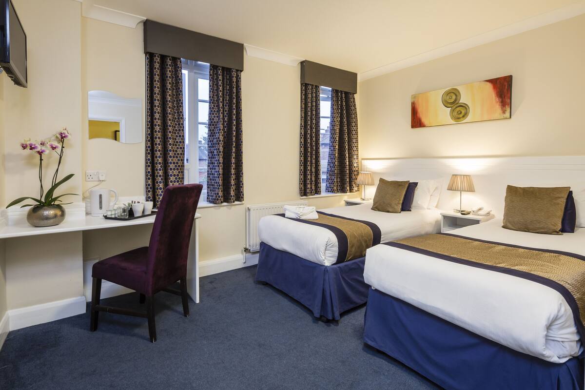 Quarto de hotel com duas camas, uma escrivaninha, uma cadeira e uma janela com cortinas.