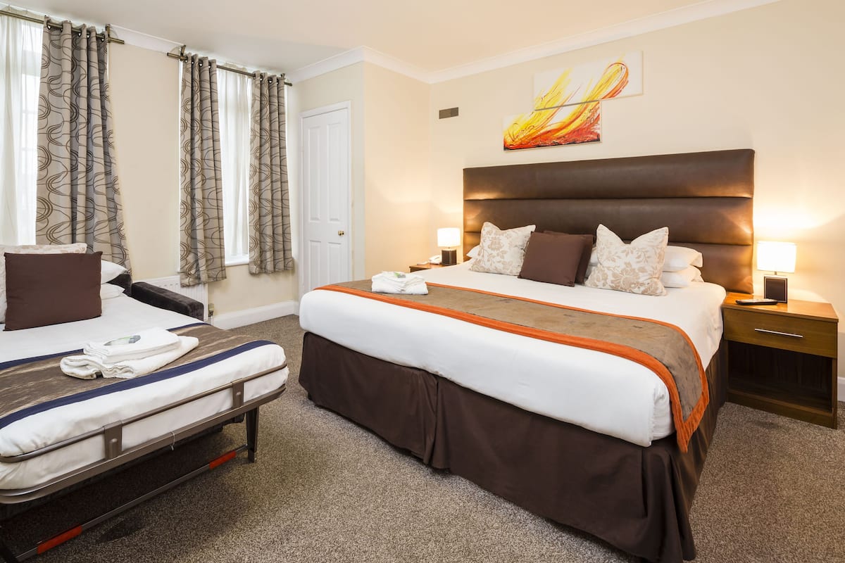 Quarto de hotel com uma cama grande, uma cama menor, duas mesinhas de cabeceira, uma mesa de apoio, uma janela com cortinas e um quadro na parede.