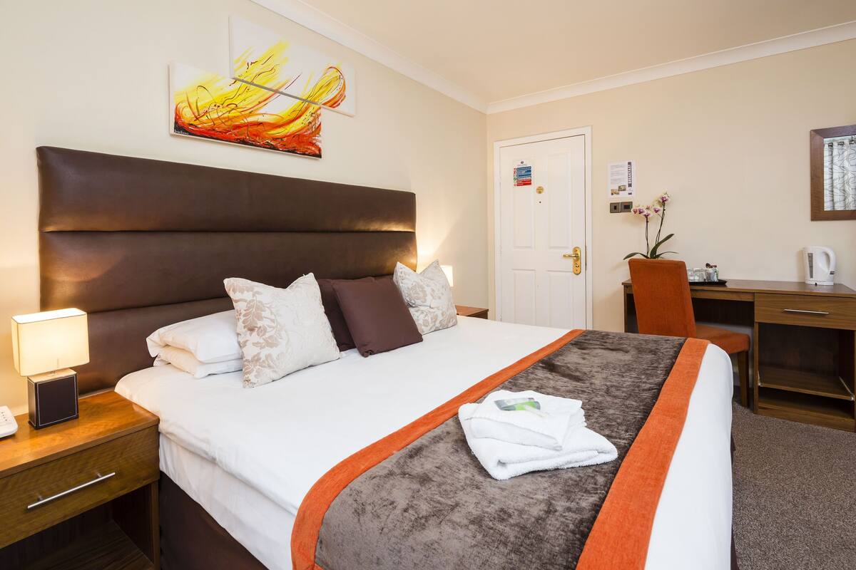 Quarto de hotel com uma cama grande, uma escrivaninha, uma cadeira, uma mesa de cabeceira com abajur e um quadro na parede.