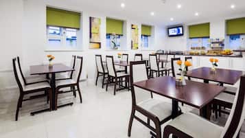 A dining area with multiple tables and chairs, a kitchen in the background, and a television mounted on the wall.