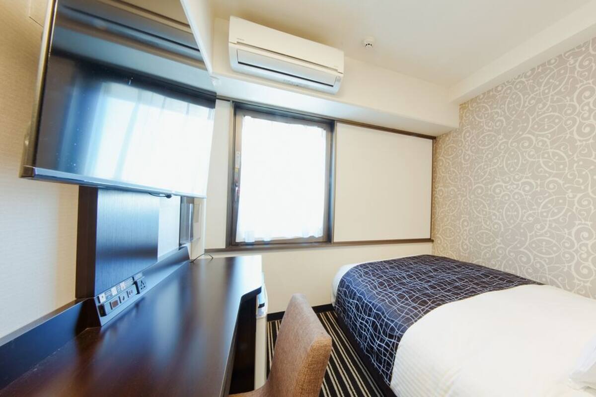 A hotel room with a bed, a desk, an air conditioning unit, and a window.