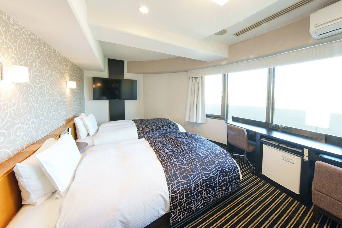 A hotel room with two beds, a TV, a minibar, and a large window with curtains.
