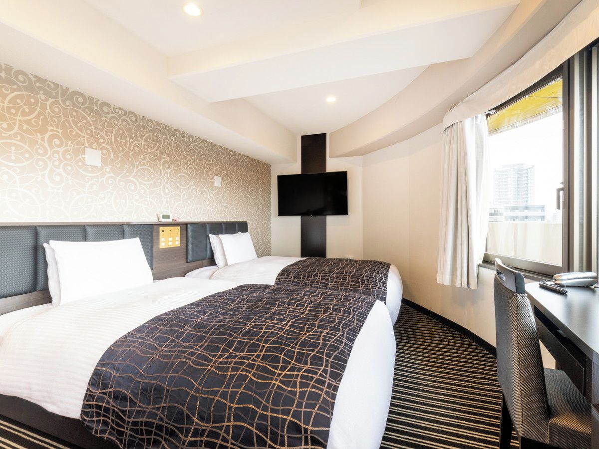A hotel room with two beds, a flat-screen TV mounted on the wall, a desk with a chair, and a window with curtains.