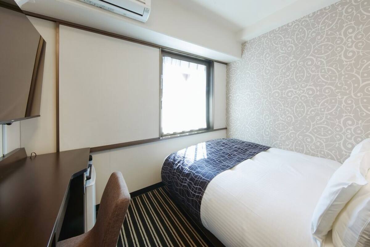 A small, compact hotel room with a bed, a desk, and a window.