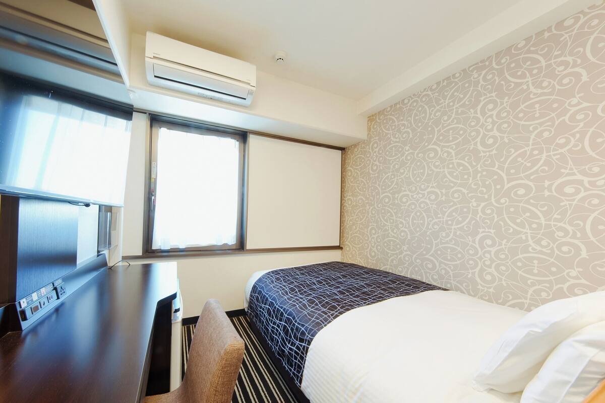 A hotel room with a bed, a desk, an air conditioning unit, a window, and patterned wallpaper.