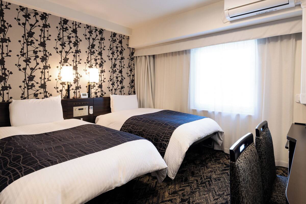A hotel room with two beds, a nightstand with a lamp, an air conditioning unit, and a window with curtains.