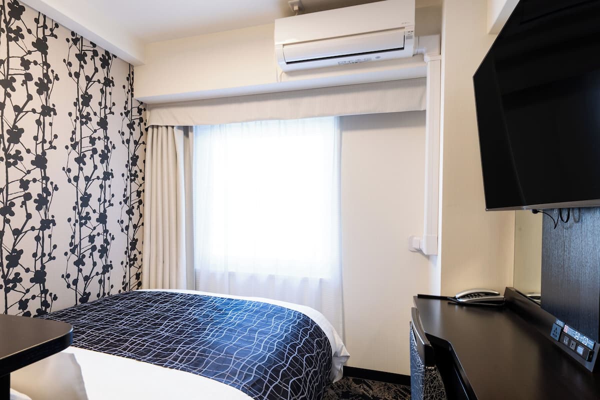 A hotel room with a bed, a desk with a telephone, an air conditioning unit, and a television.