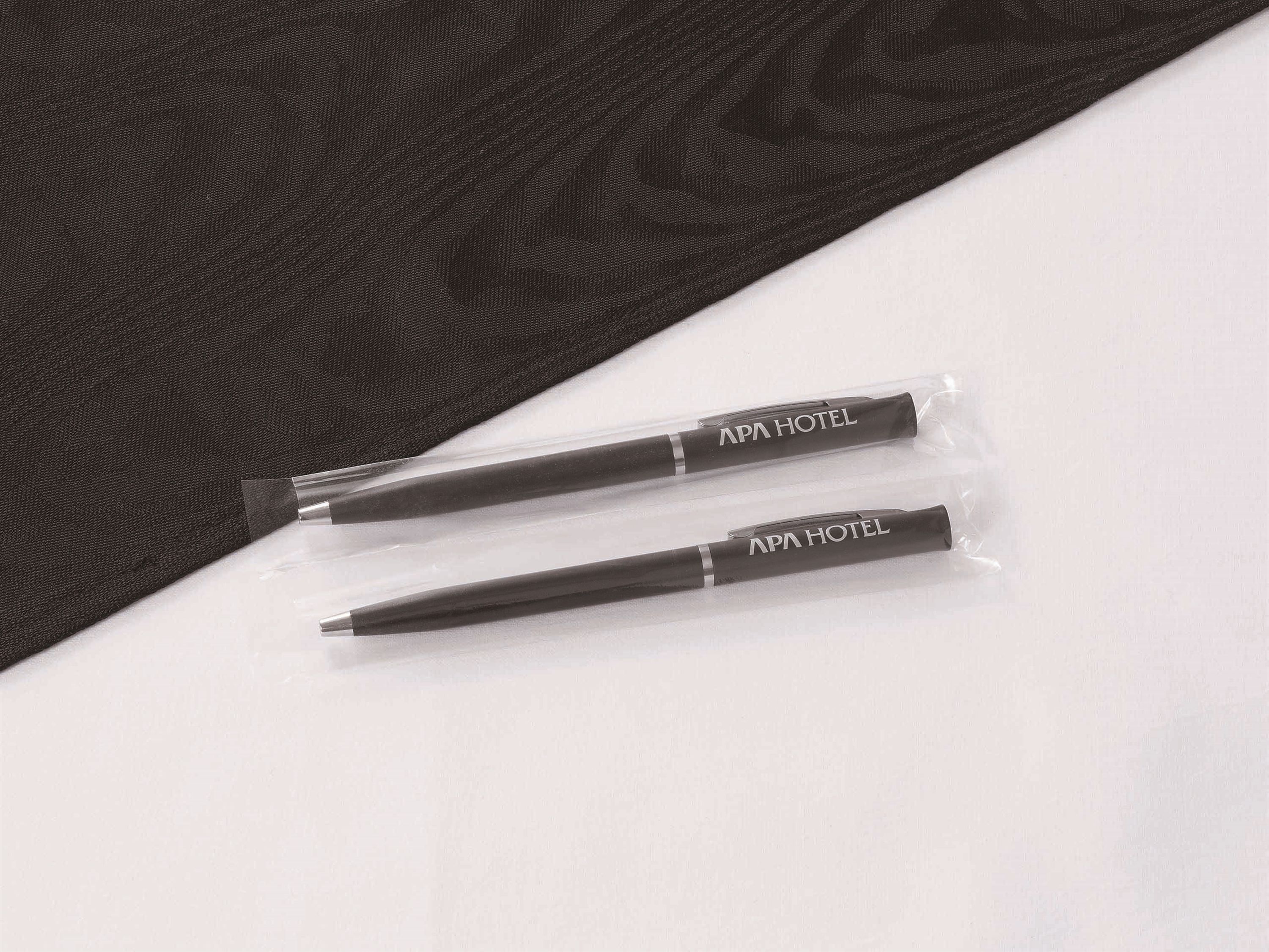 Two pens with "APA HOTEL" branding in clear plastic packaging, placed on a white surface with a dark fabric background.