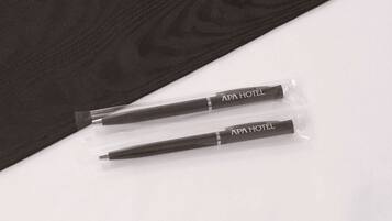 Two pens with "APA HOTEL" branding in clear plastic packaging, placed on a white surface with a dark fabric background.
