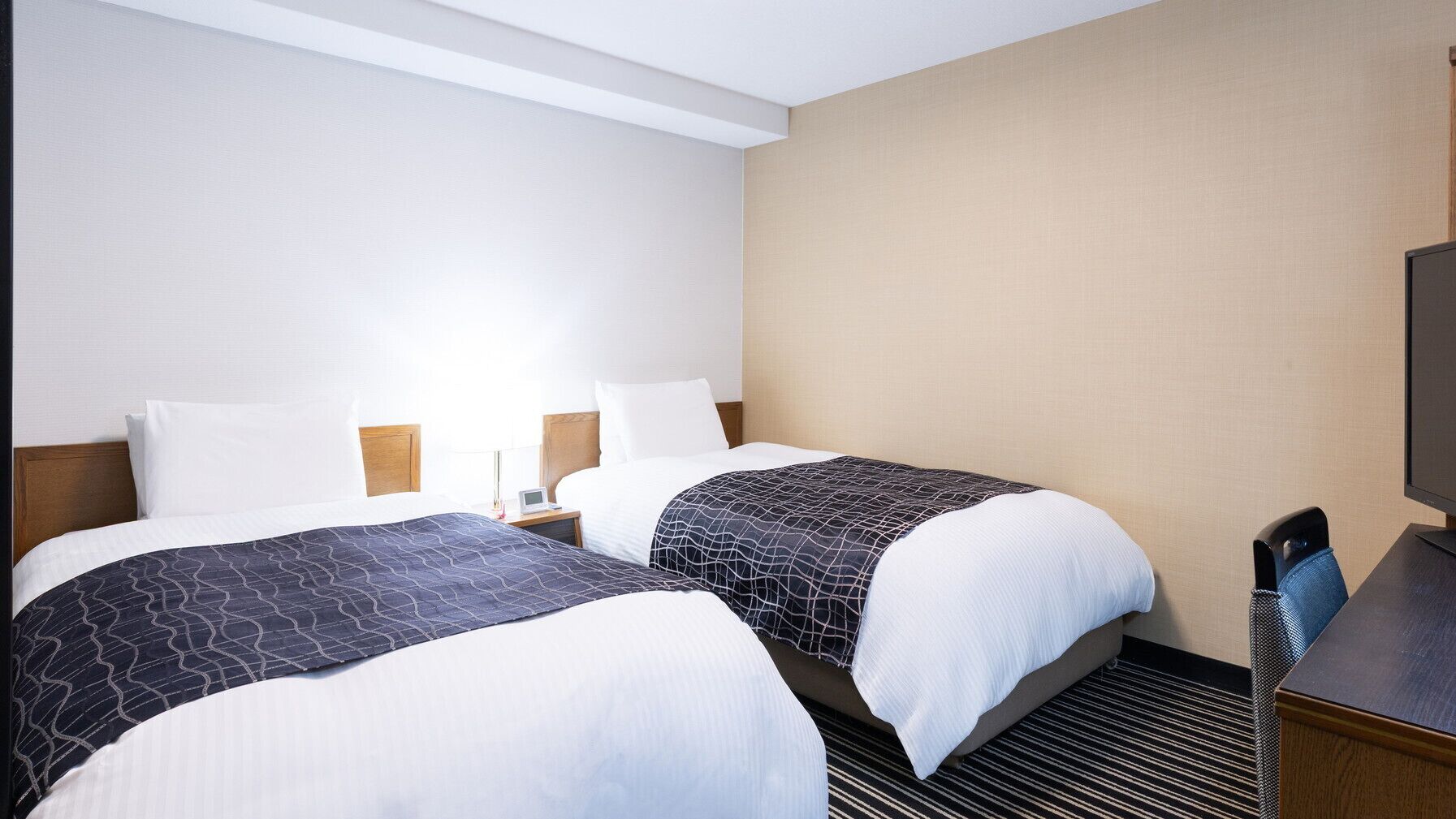 A hotel room with two beds, a desk, a chair, a TV, and a lamp.