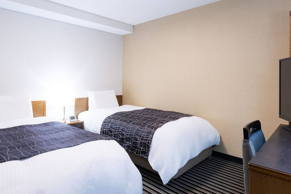 A hotel room with two beds, a desk, a chair, a TV, and a lamp.