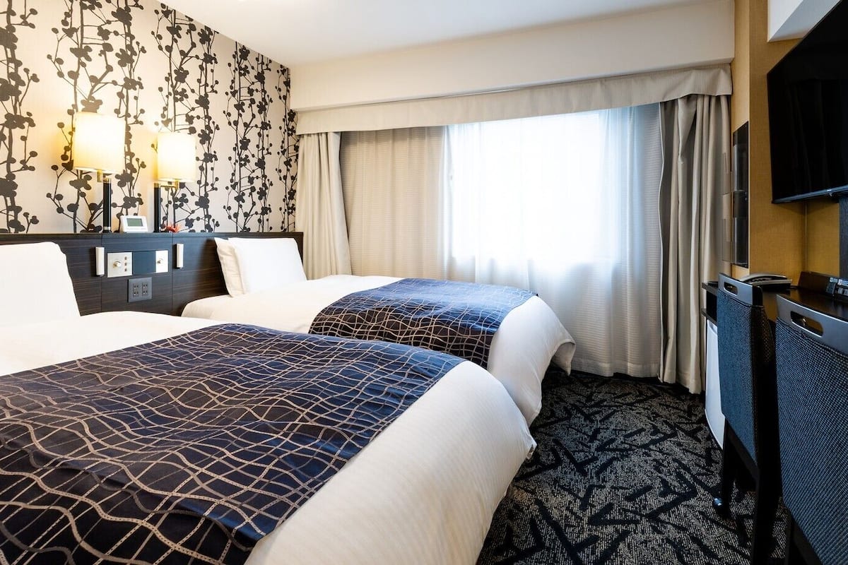 A hotel room with two beds, a flat-screen TV, a desk, and a large window with curtains.