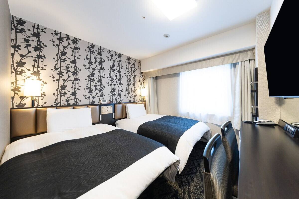 A hotel room with two beds, a desk with a flat-screen TV, a telephone, and a wall-mounted lamp.