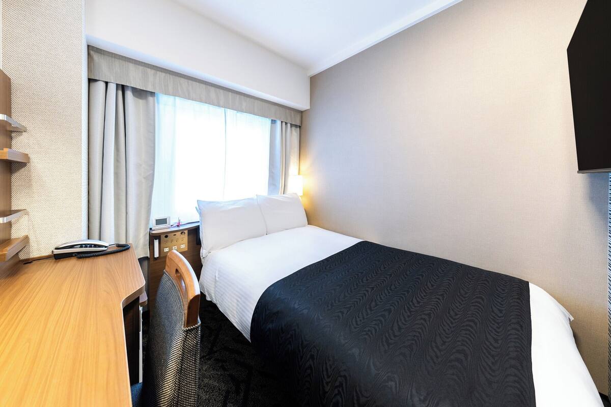 A hotel room with a bed, a desk, a chair, a telephone, and a window with curtains.