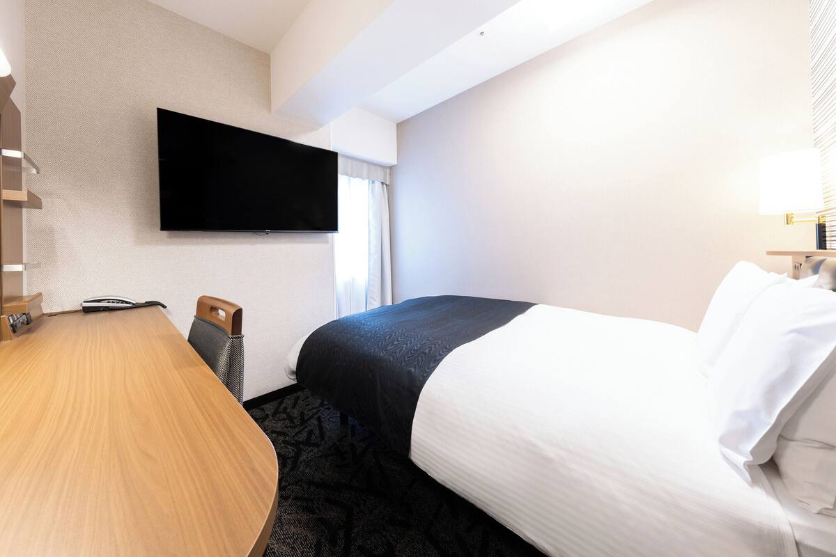A hotel room with a bed, a desk, a flat-screen TV mounted on the wall, and a window with curtains.