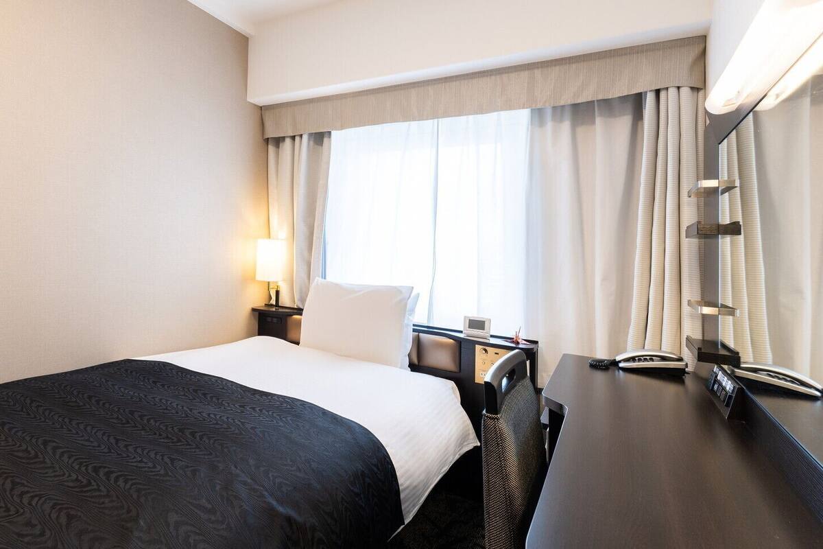 A hotel room with a bed, bedside table, lamp, and a desk with a telephone.