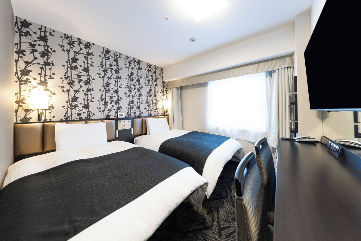 A hotel room with two beds, a desk with a flat-screen TV, and a window with curtains.