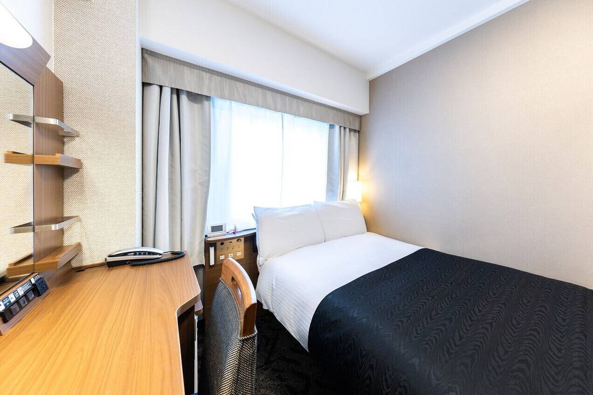 A hotel room with a bed, a desk with a telephone, a chair, a nightstand, and a window with curtains.