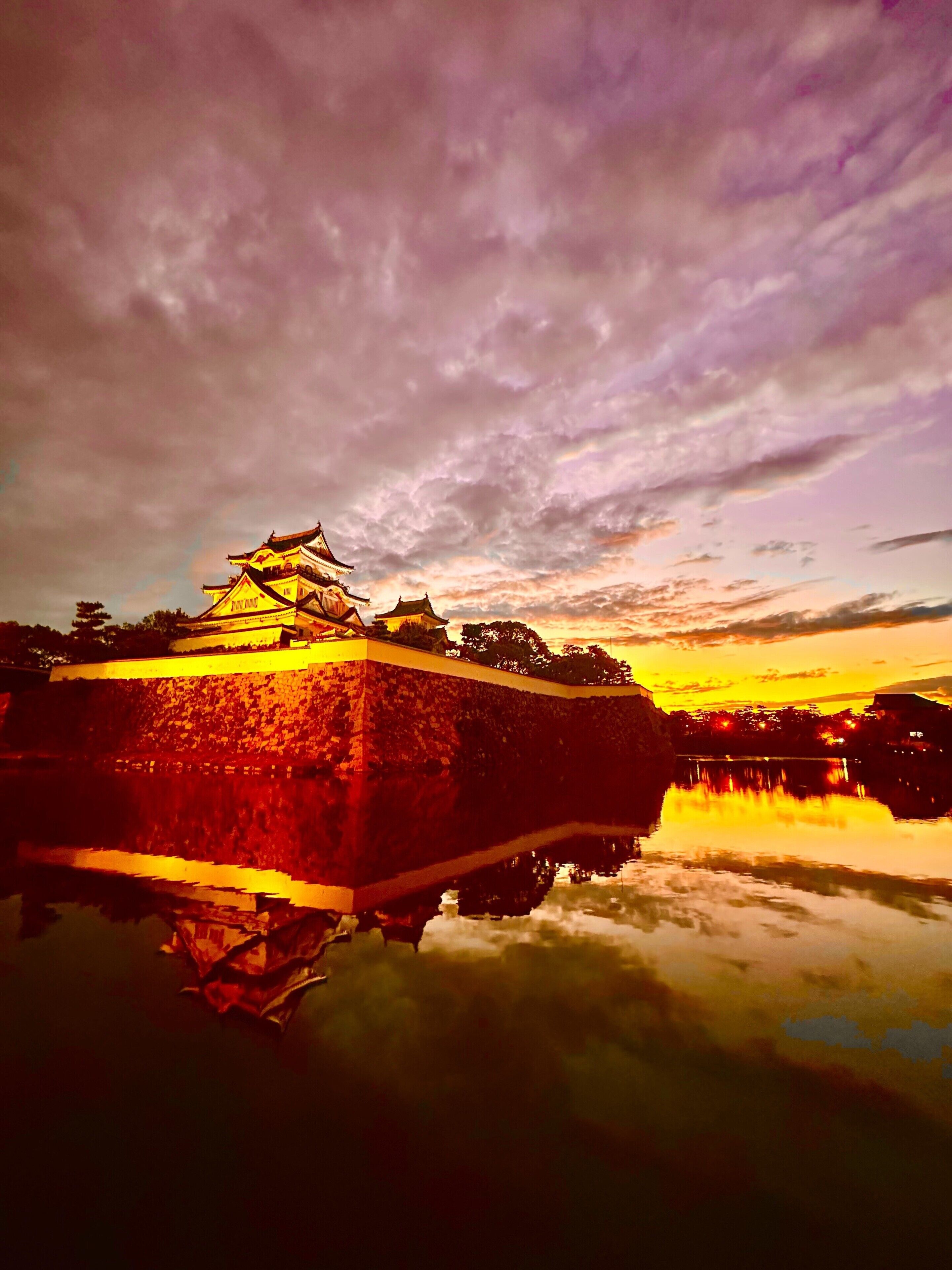 A traditional Japanese castle with a stone wall and reflection in water, under a cloudy sky.