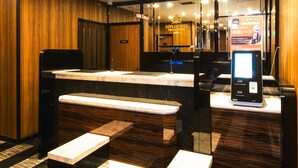 Zona de recepción de un hotel con mostrador de mármol, quiosco de registro digital y un espejo que refleja el interior.