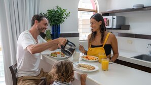 Una família dinant al taulell de cuina, amb un microones, una planta en test i una finestra amb cortines.