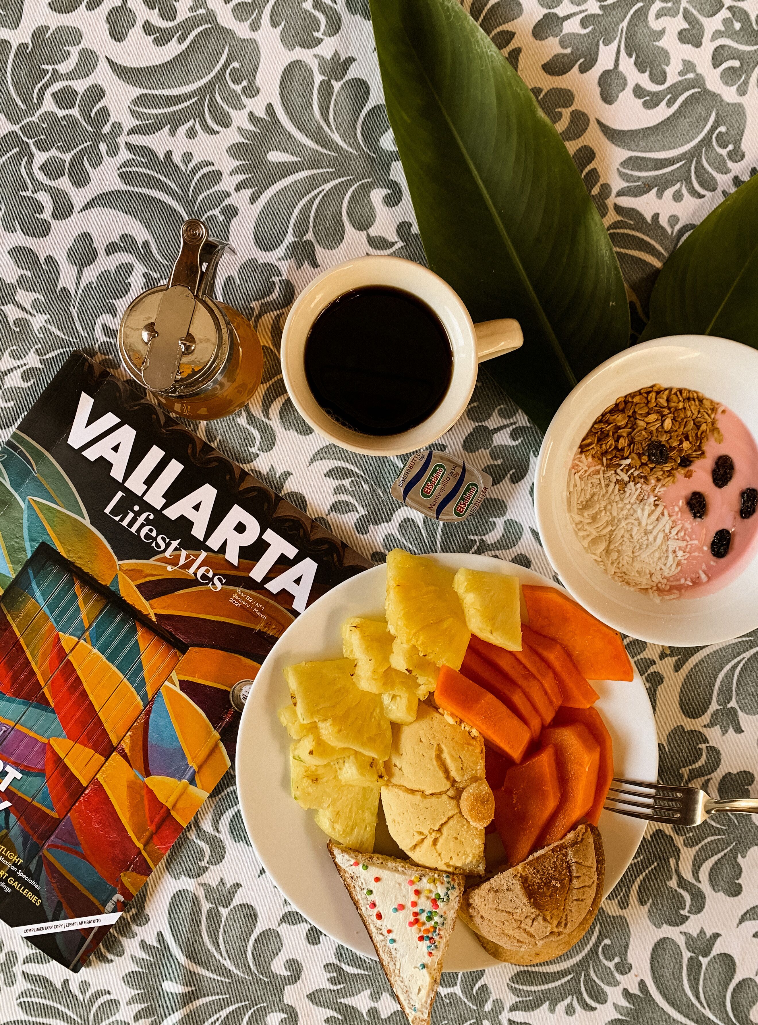 A table with a patterned tablecloth, a cup of coffee, a plate with pineapple chunks, carrot slices, and a slice of bread with colorful sprinkles.
