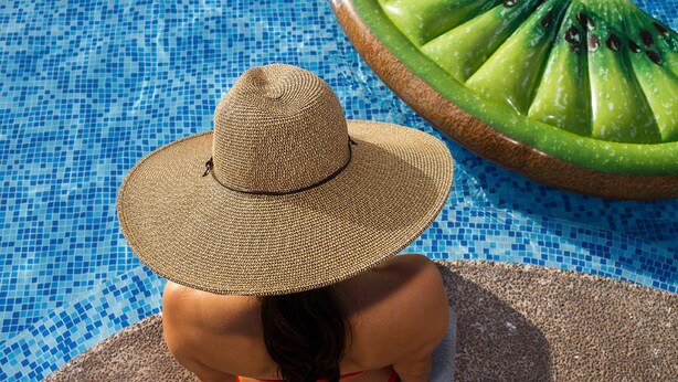 Una persona asseguda al costat d'una piscina, portant un barret gran i un banyador vermell.