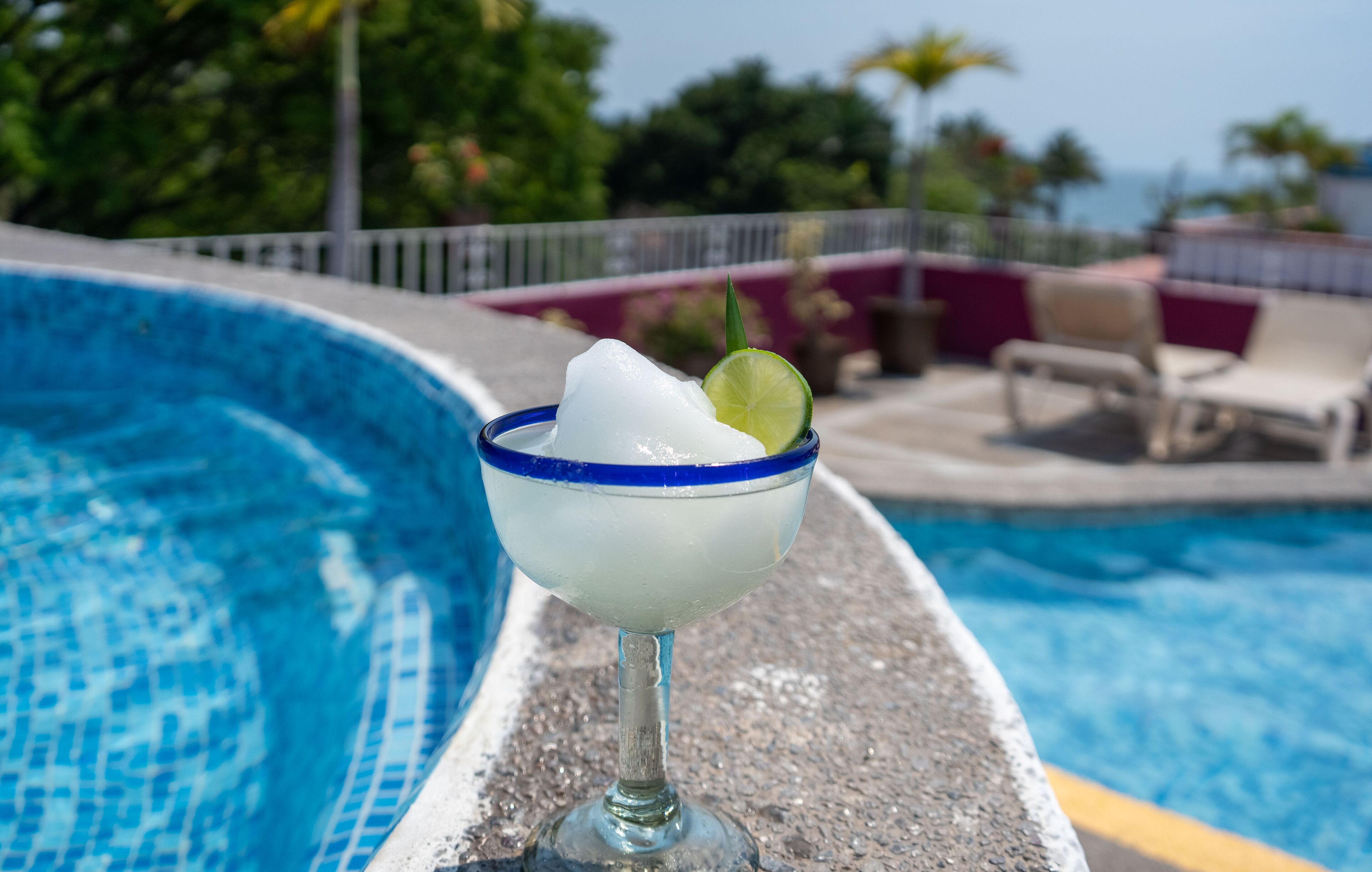 A cocktail in a blue-rimmed glass with an ice cube and a lime slice, placed on a poolside ledge.